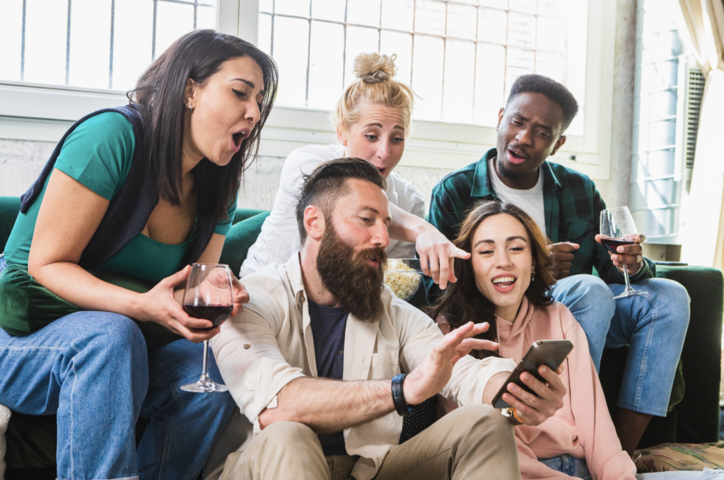 A group of friends sitting together on a couch, sharing a fun moment while looking at a phone screen. They are casually enjoying snacks and drinks, reacting enthusiastically to something on the phone. The image captures a relaxed, social atmosphere, highlighting connection, enjoyment, and group interaction.